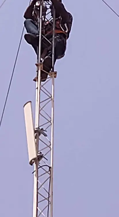 close-up, upward-looking shot of a technician scaling a telecommunications tower. The person is wearing a safety harness and is positioned high up on the steel lattice structure